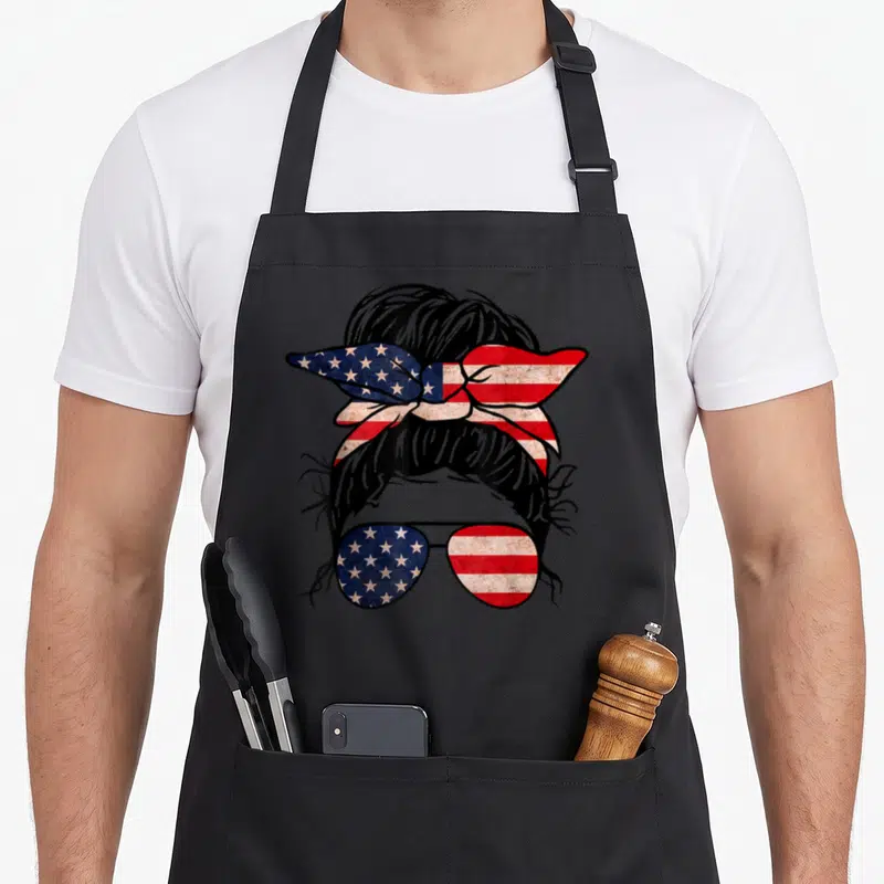 Messy 4th of july apron - cartoon messy bun with red white blue flag headband and star-spangled sunglasses — shown on a black cotton canvas apron against a white background
