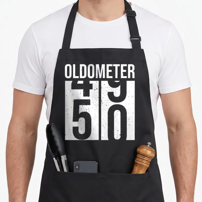 50th born day apron - Oldometer text with 49 and 50 numbers in white distressed rectangles on grey background — shown on a black cotton canvas apron against a white background