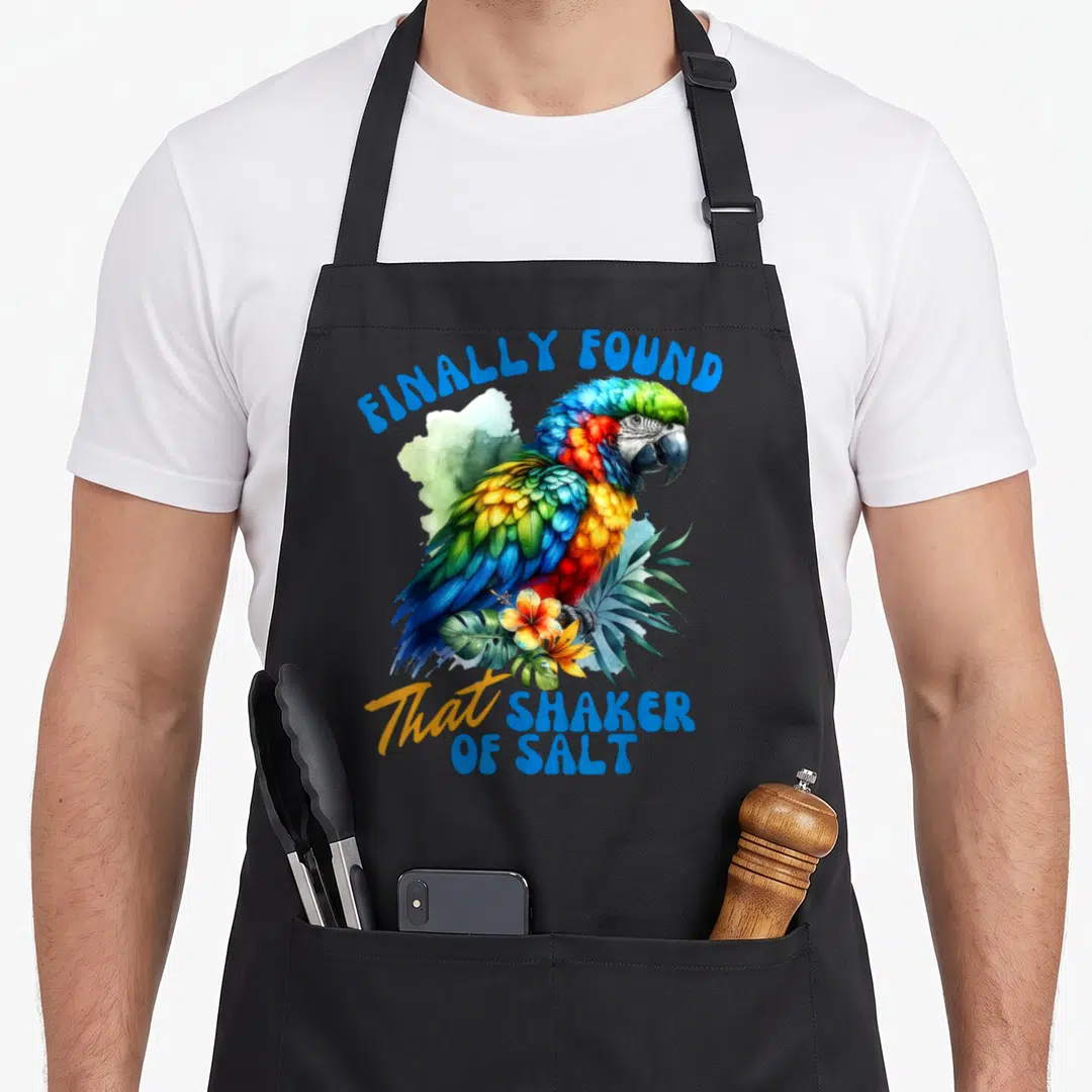 Finally bird apron - Colorful parrot with FINALLY FOUND That SHAKER OF SALT text and tropical watercolor foliage — shown on a black cotton canvas apron against a white background