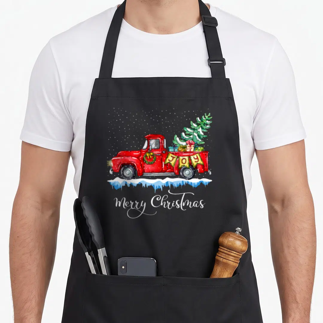 Christmas old fashioned aprons - Red truck carrying tree and gifts with JOY banner and Merry Christmas text below — shown on a black cotton canvas apron against a white background