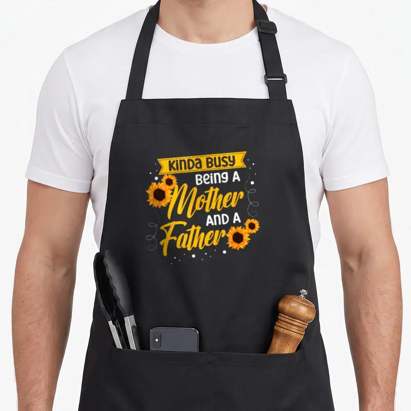 Kinda mother's apron - KINDA BUSY Being A Mother AND A Father text with sunflowers and yellow ribbon — shown on a black cotton canvas apron against a white background