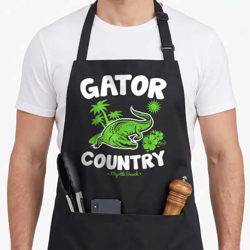 Myrtle country apron - Green cartoon alligator with palm trees and Gator Country text — shown on a black cotton canvas apron against a white background
