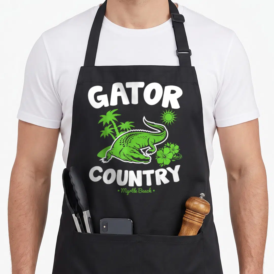 Myrtle country apron - Green cartoon alligator with palm trees and Gator Country text — shown on a black cotton canvas apron against a white background