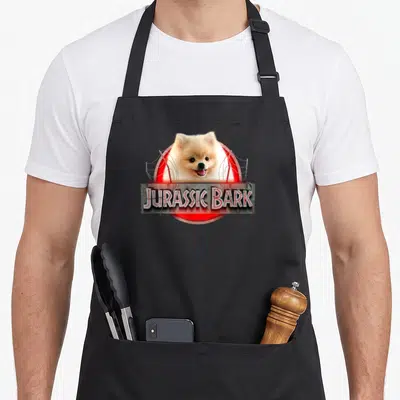 Jurassic cooking with dog apron - Pomeranian head in red circle with Jurassic Bark stone plaque and wire scratches — shown on a black cotton canvas apron against a white background