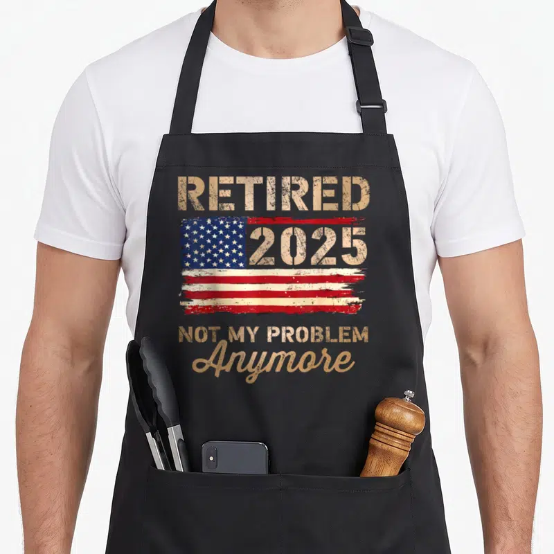 Flag retirement apron - Distressed USA flag with Retired 2025 Not My Problem Anymore text in gold and red — shown on a black cotton canvas apron against a white background