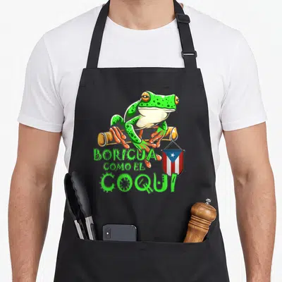 Rican frog aprons - Cartoon frog perched on branch with BORICUA COMO EL COQUI text and Puerto Rican flag — shown on a black cotton canvas apron against a white background