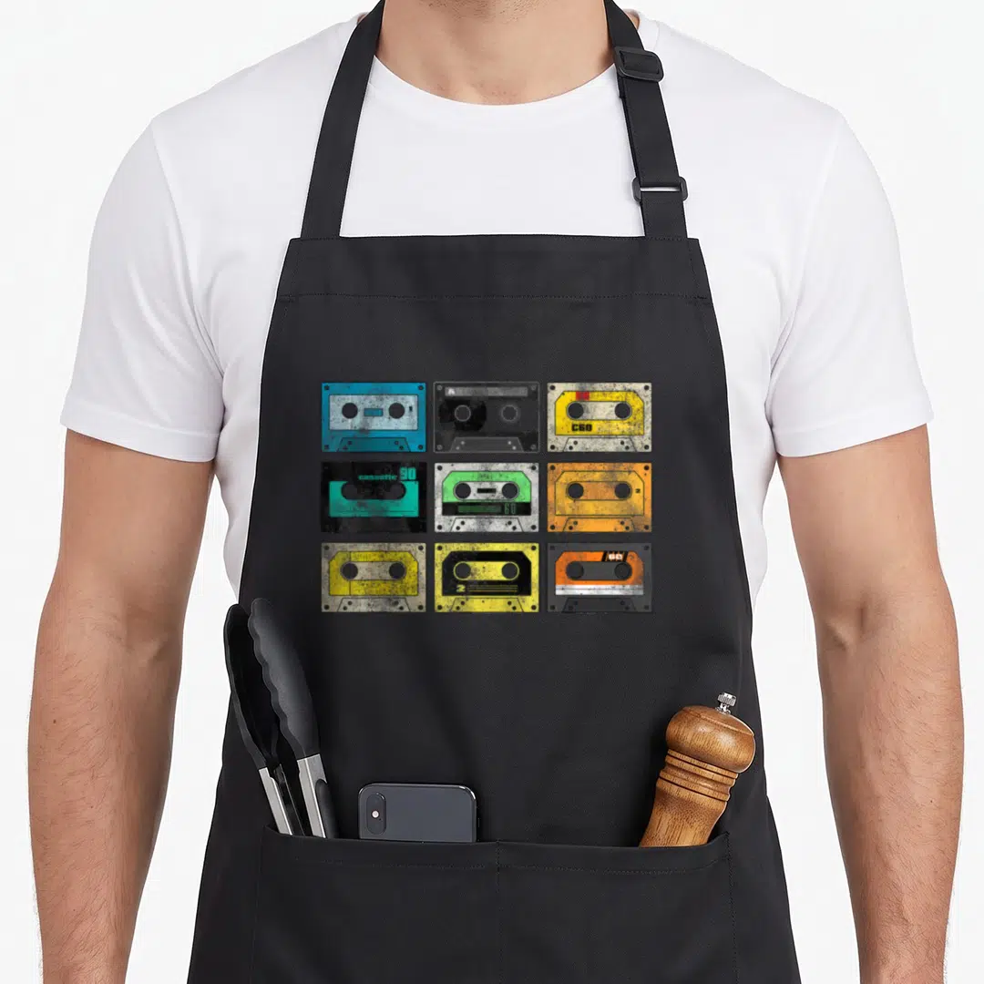 Audio 80s apron - Nine colorful cassette tapes in a 3x3 grid showing C60 and stereo labels — shown on a black cotton canvas apron against a white background