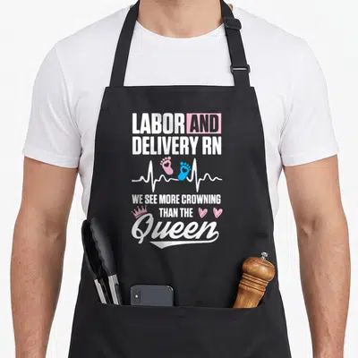 Crowning nurse apron - Labor and Delivery RN text with heartbeat line, pink and blue footprints, crown, and hearts — shown on a black cotton canvas apron against a white background