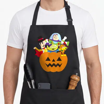 Funny halloween pumpkin apron - Orange pumpkin with cowboy astronaut alien and dog popping from behind in cartoon style — shown on a black cotton canvas apron against a white background