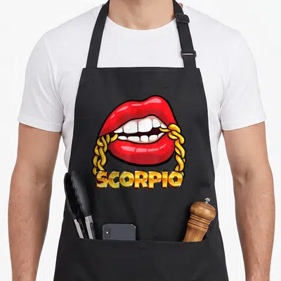 Juicy zodiac aprons - Red cartoon lips biting gold chain framing SCORPIO text — shown on a black cotton canvas apron against a white background
