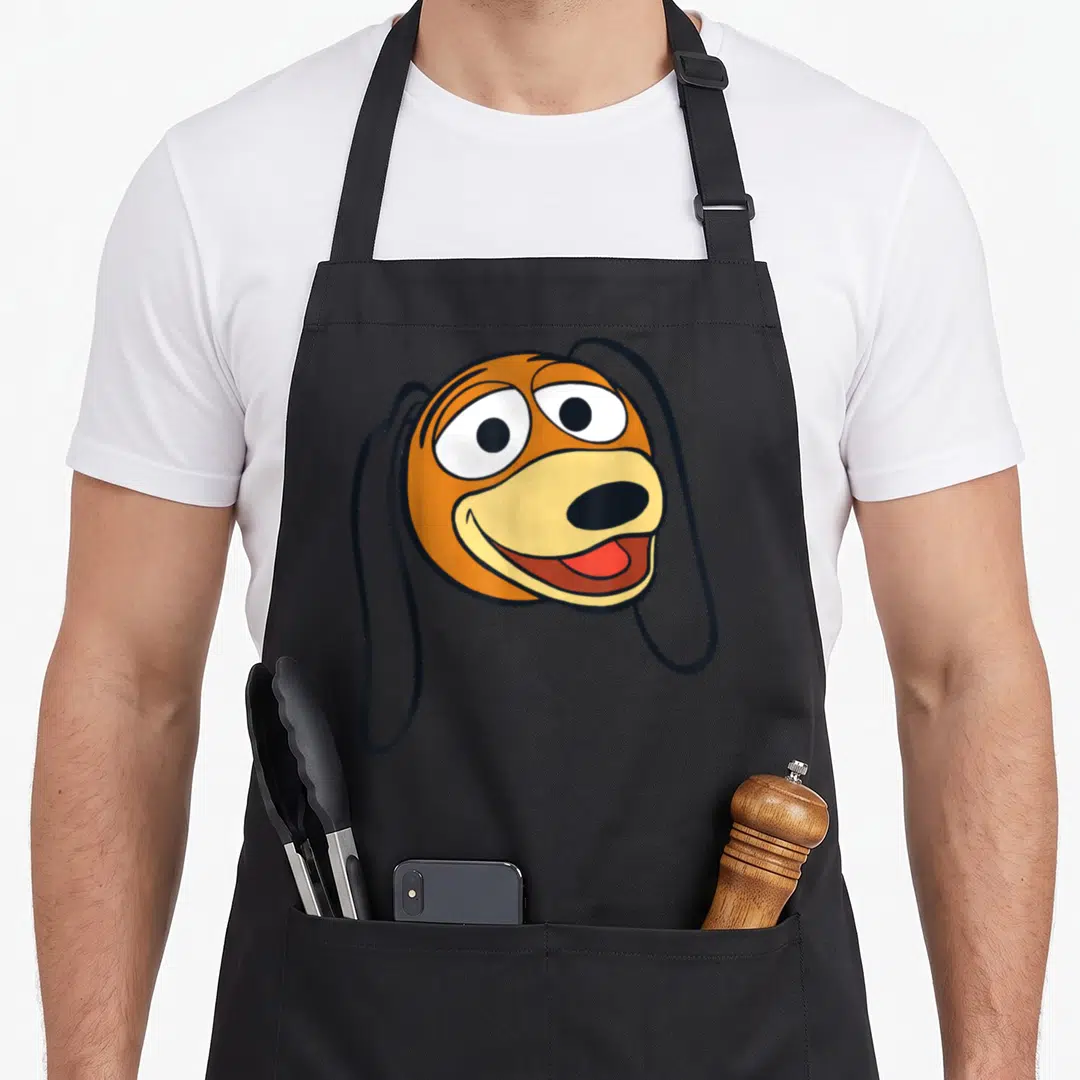 Funny slinky dog apron - Cartoon dog face in orange and yellow with long black ears — shown on a black cotton canvas apron against a white background