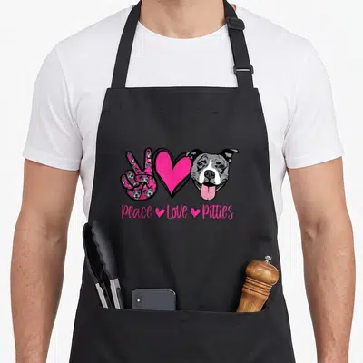 Pit dog apron - Peace Love Pitties text under pink peace sign, heart, and grey cartoon dog head — shown on a black cotton canvas apron against a white background