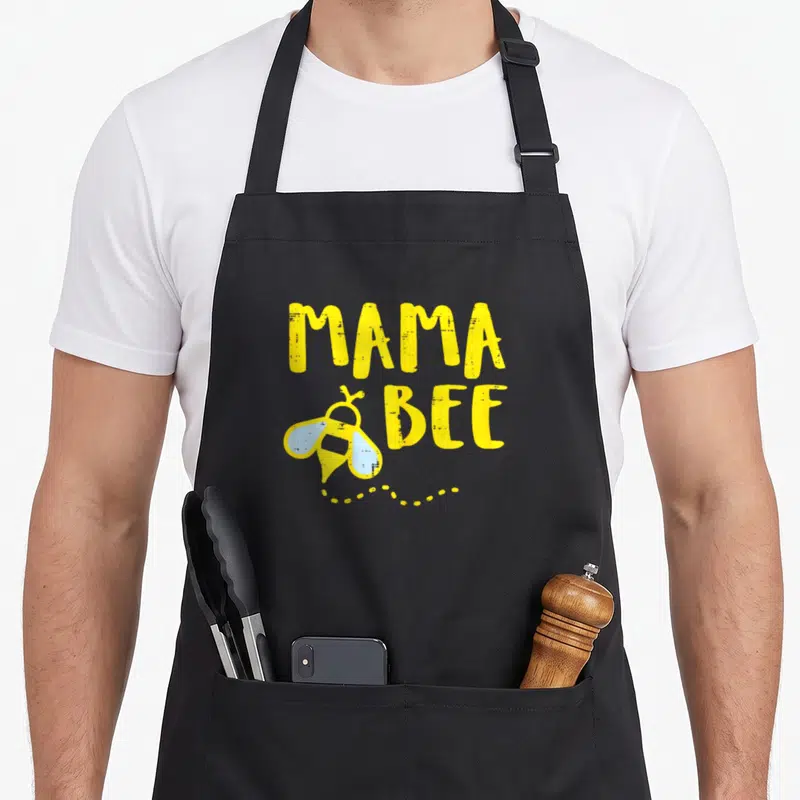 Bee mama apron - MAMA BEE text with yellow cartoon bee and light blue wings above a dotted trail — shown on a black cotton canvas apron against a white background