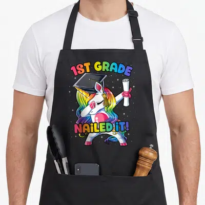 Class unicorn apron - Dabbing white unicorn with rainbow mane holding diploma under 1ST GRADE NAILED IT! text — shown on a black cotton canvas apron against a white background