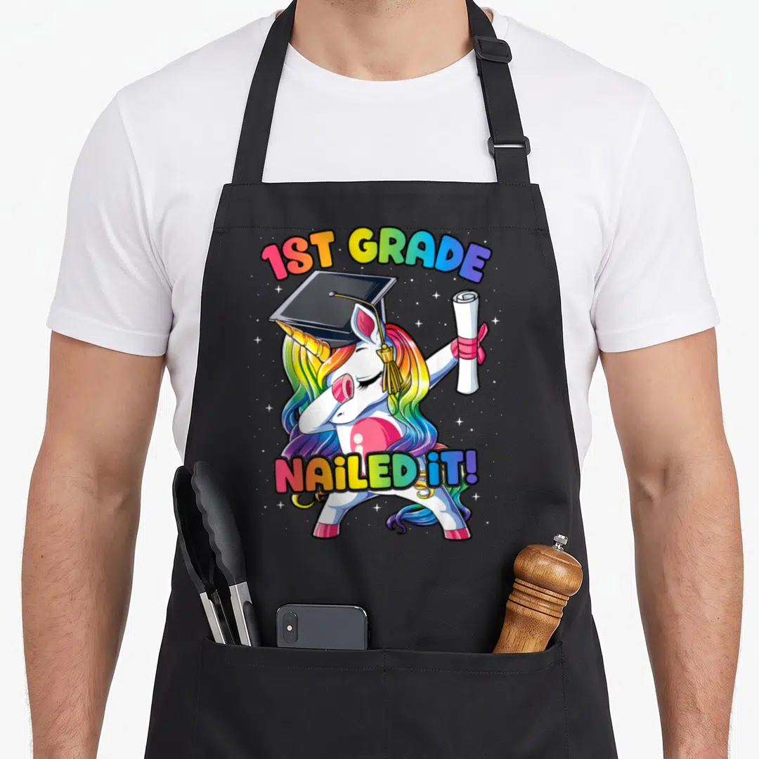 Class unicorn apron - Dabbing white unicorn with rainbow mane holding diploma under 1ST GRADE NAILED IT! text — shown on a black cotton canvas apron against a white background