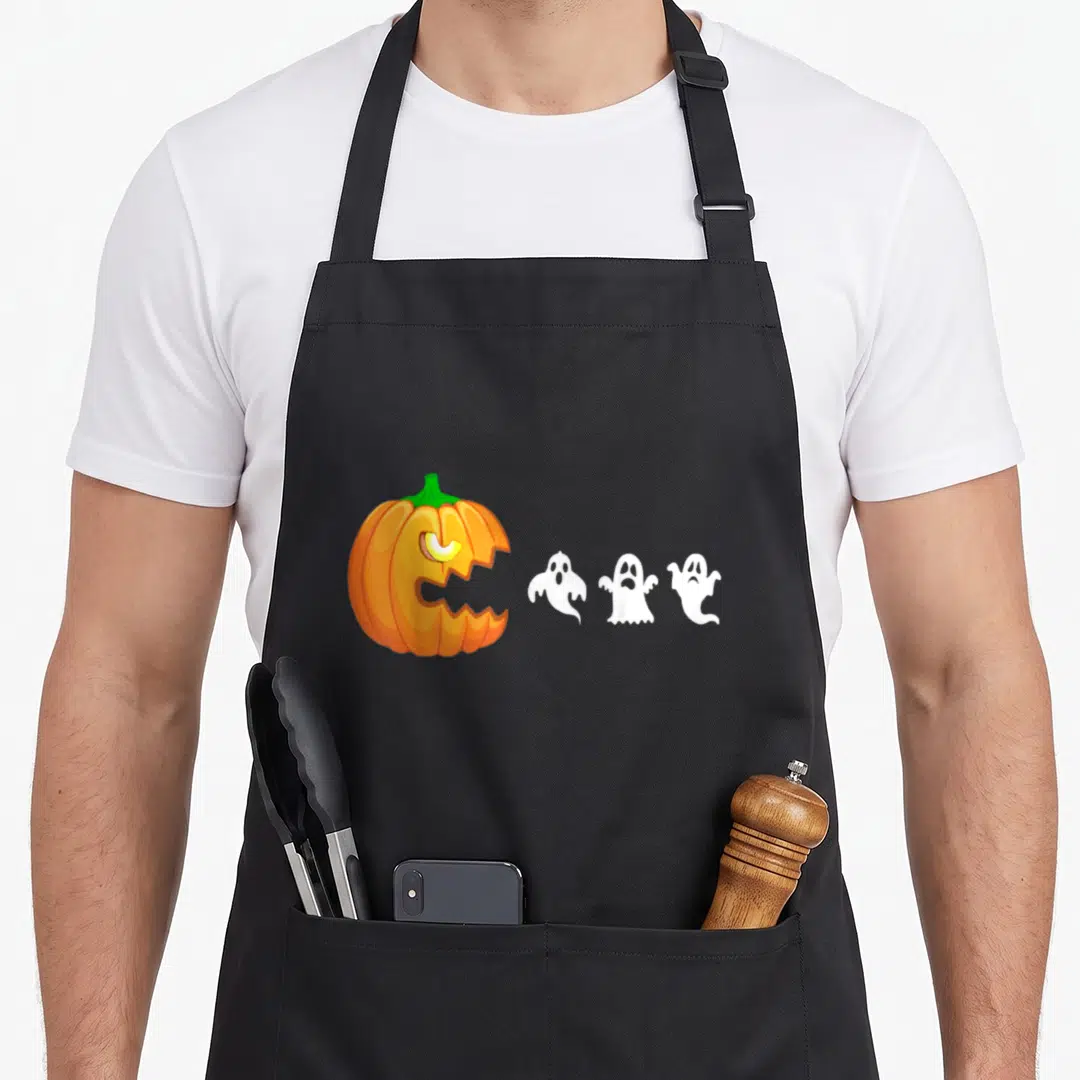 Eating pumpkin apron - Cartoon orange jack-o-lantern with green stem and three white ghosts in a row — shown on a black cotton canvas apron against a white background