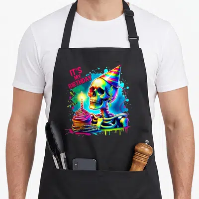 Cake funny halloween apron - Rainbow skeleton holding cupcake with IT'S MY BIRTHDAY text and neon splatters — shown on a black cotton canvas apron against a white background