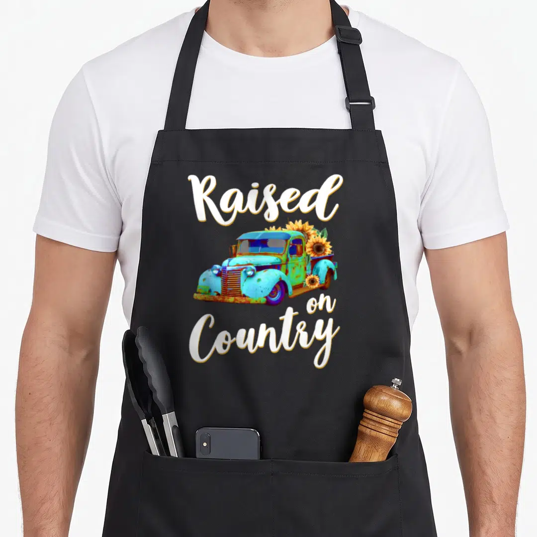 Raised truck apron - Vintage blue truck with sunflowers and Raised on Country text in white script — shown on a black cotton canvas apron against a white background