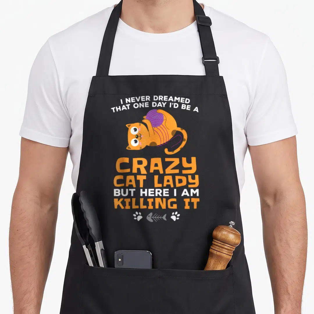 Dreamed crazy cat lady apron - Orange cartoon cat holding purple yarn with split white and orange text and bottom paw — shown on a black cotton canvas apron against a white background