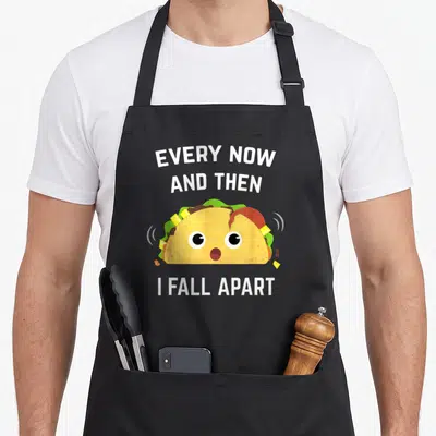 Then taco apron - Cartoon taco splitting open with white text reading Every Now and Then I Fall Apart — shown on a black cotton canvas apron against a white background