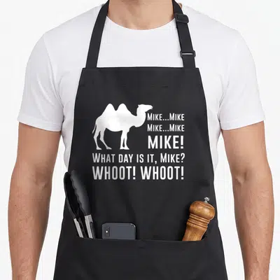 Funny hump camel apron - White camel silhouette next to stacked text reading Mike What Day Is It Whoot Whoot — shown on a black cotton canvas apron against a white background