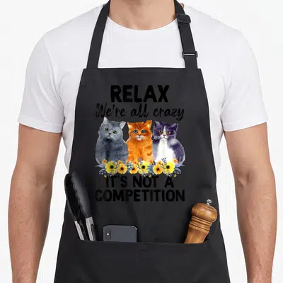 Relax black cat aprons - Three watercolor cats in grey orange and purple behind yellow flowers with humorous text — shown on a black cotton canvas apron against a white background