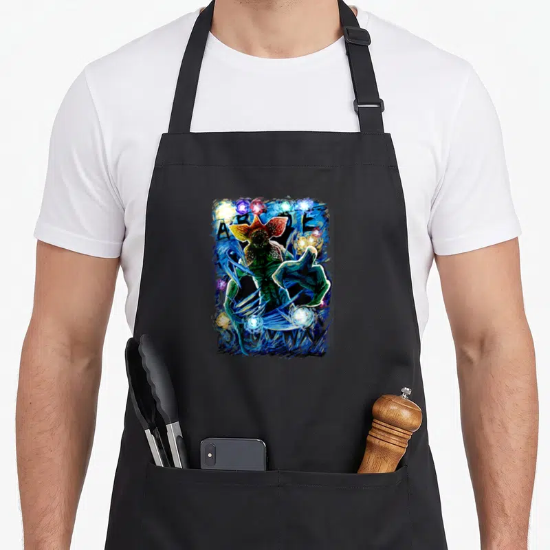 Letter starry night apron - Cartoon monster surrounded by blue energy streaks and multicolored spheres in green red — shown on a black cotton canvas apron against a white background