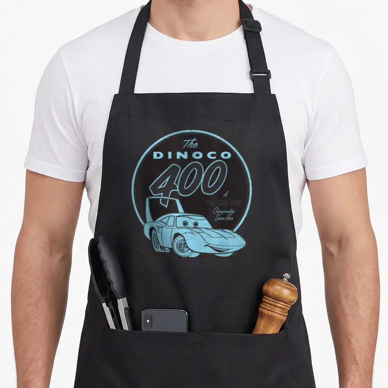 Cup car apron - Dinoco 400 Piston Cup logo with light blue cartoon race car on dark grey circle — shown on a black cotton canvas apron against a white background