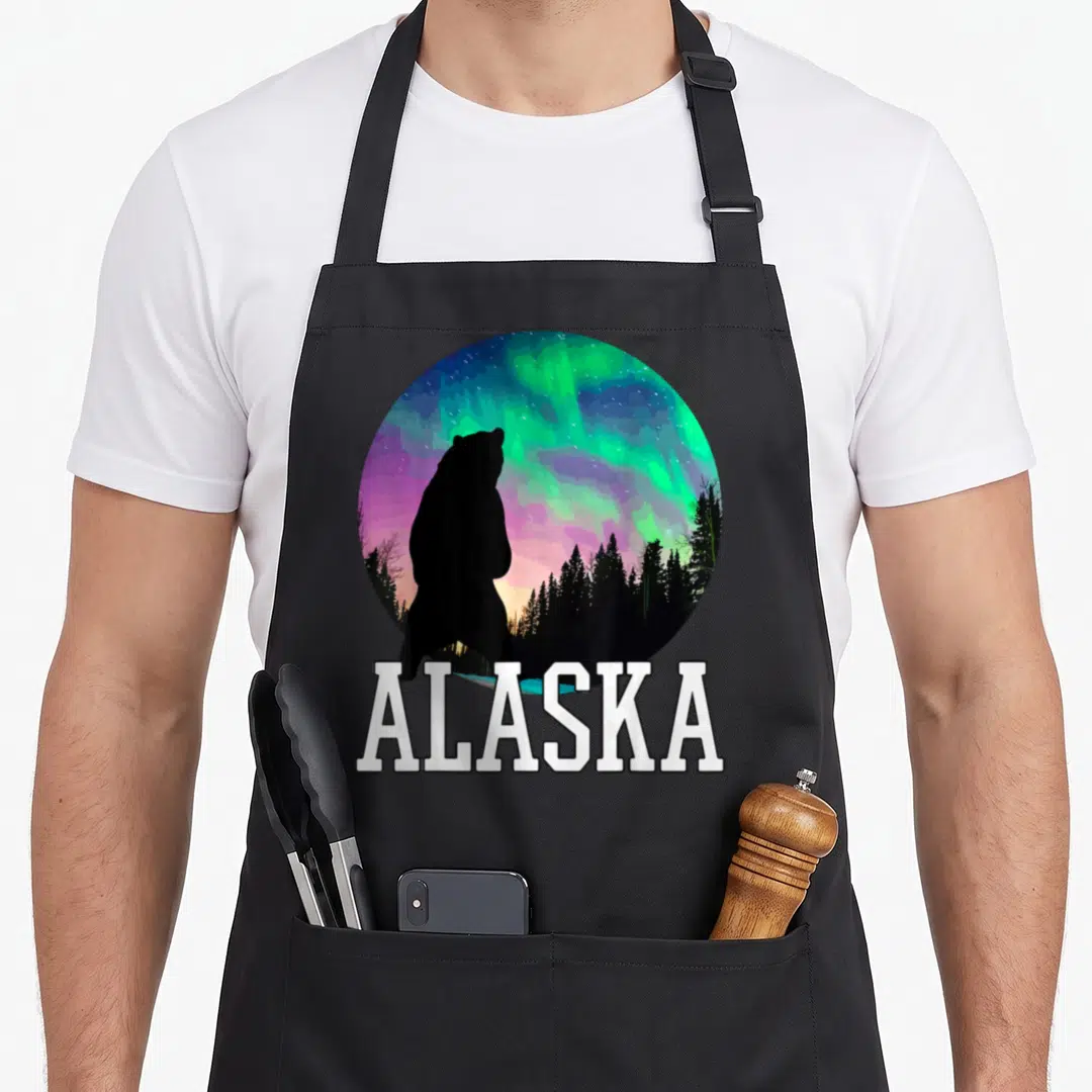 Alaska holiday apron - Bear silhouette under green purple aurora borealis with white ALASKA text below circle — shown on a black cotton canvas apron against a white background
