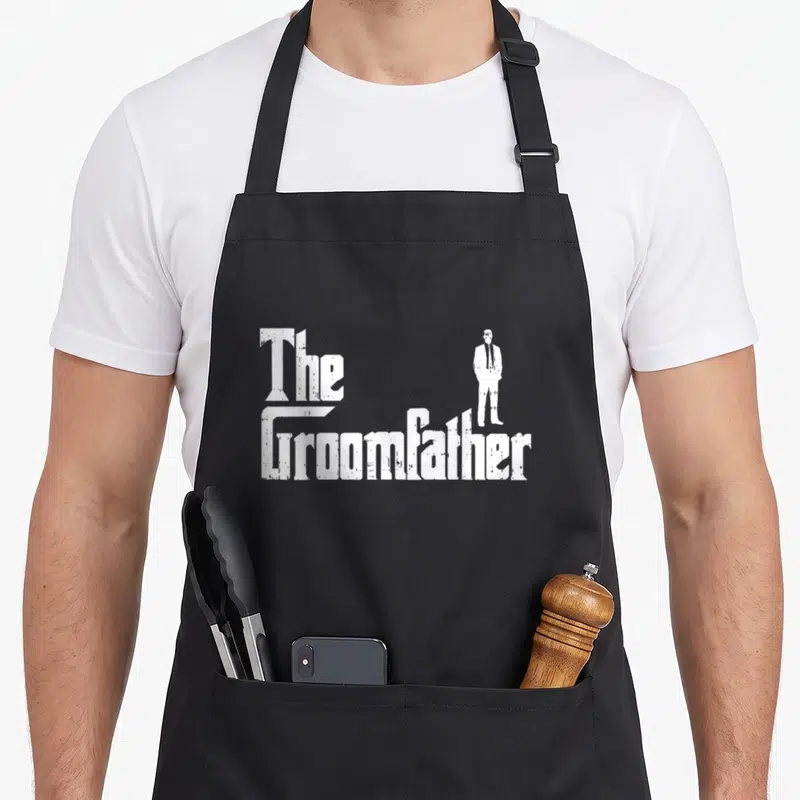 Groom funny wedding aprons - White distressed The Groomfather text with standing suit silhouette — shown on a black cotton canvas apron against a white background