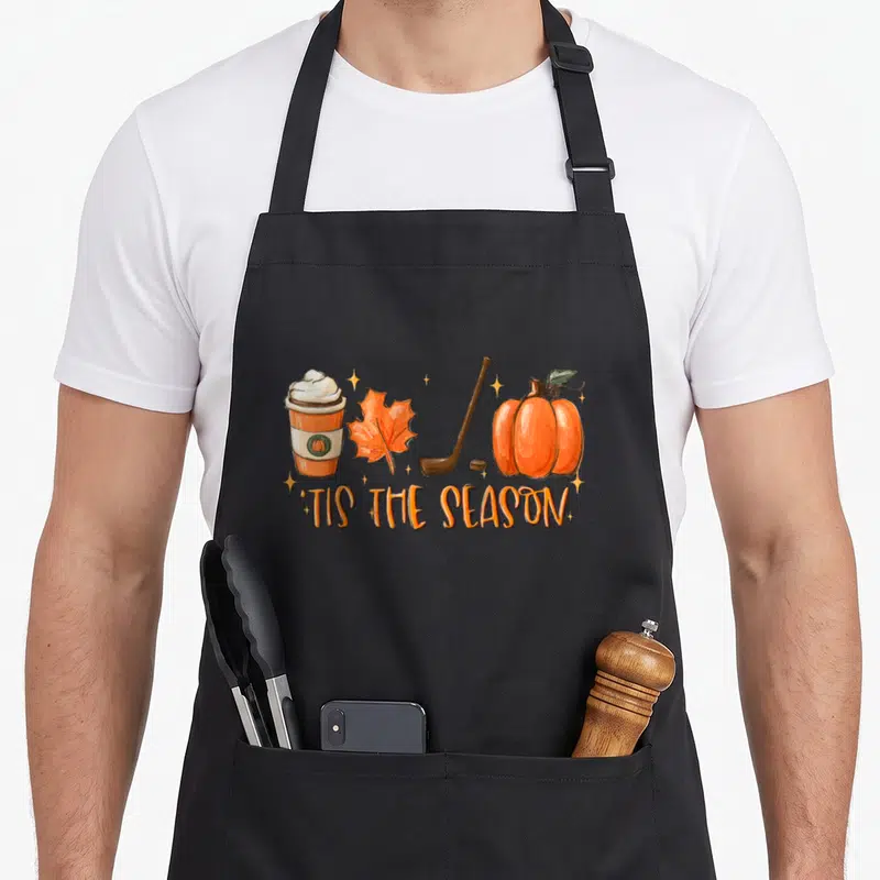 Tis halloween apron - Cartoon row of coffee cup, orange leaf, hockey stick, puck, and pumpkin above orange text — shown on a black cotton canvas apron against a white background