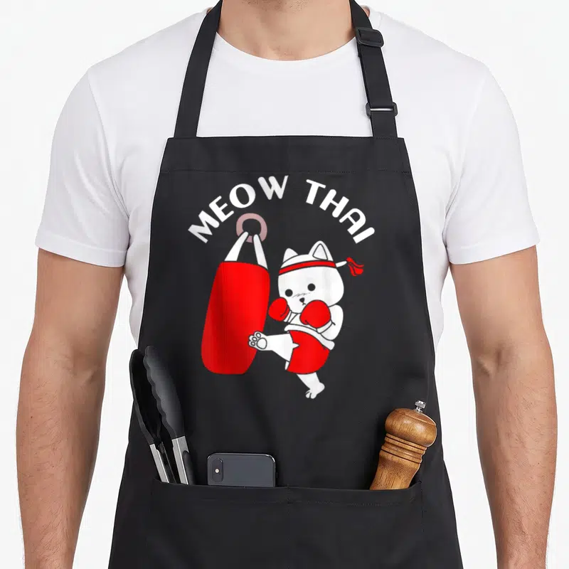 Muay cat apron - White cartoon cat kicking red bag under arched MEOW THAI text — shown on a black cotton canvas apron against a white background