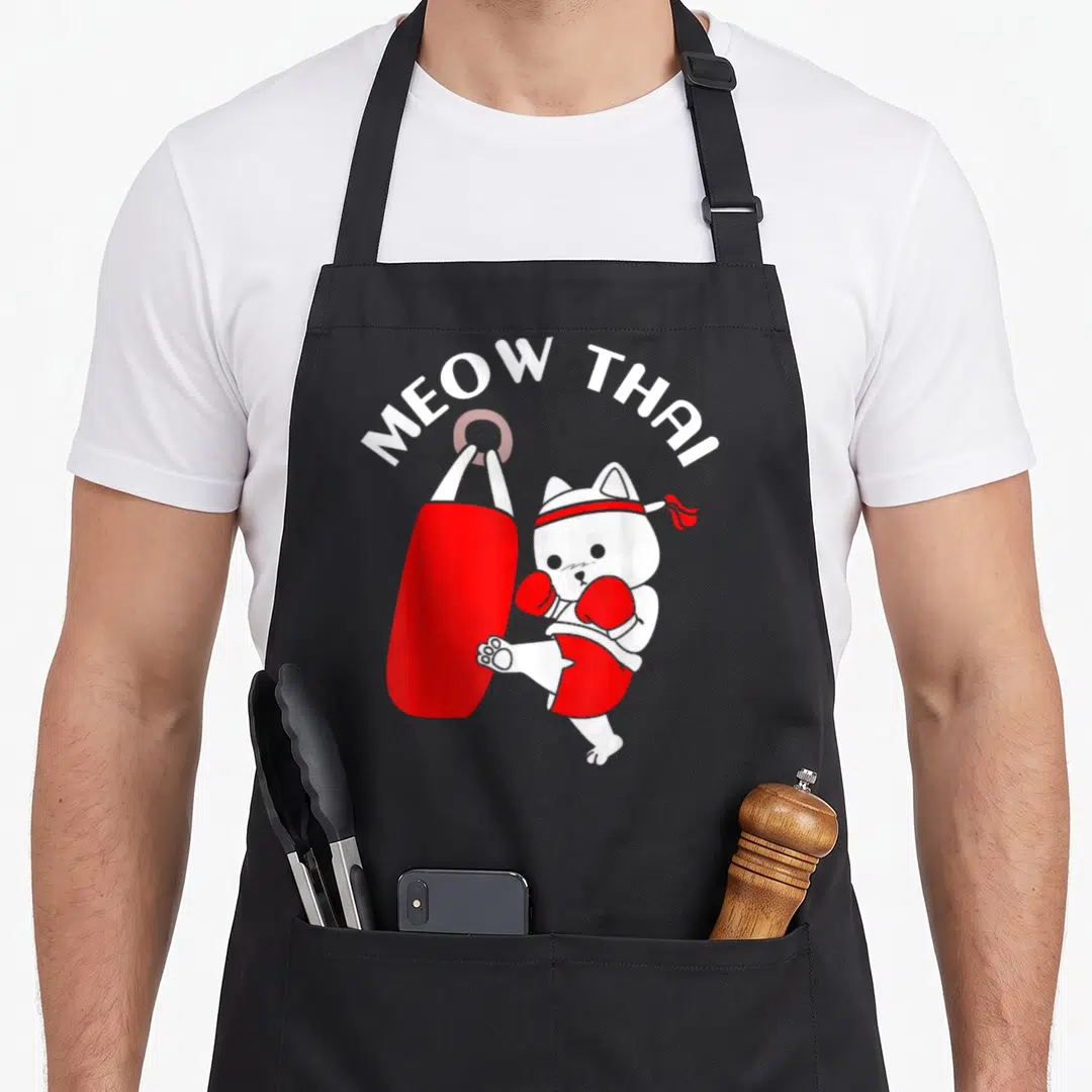 Muay cat apron - White cartoon cat kicking red bag under arched MEOW THAI text — shown on a black cotton canvas apron against a white background