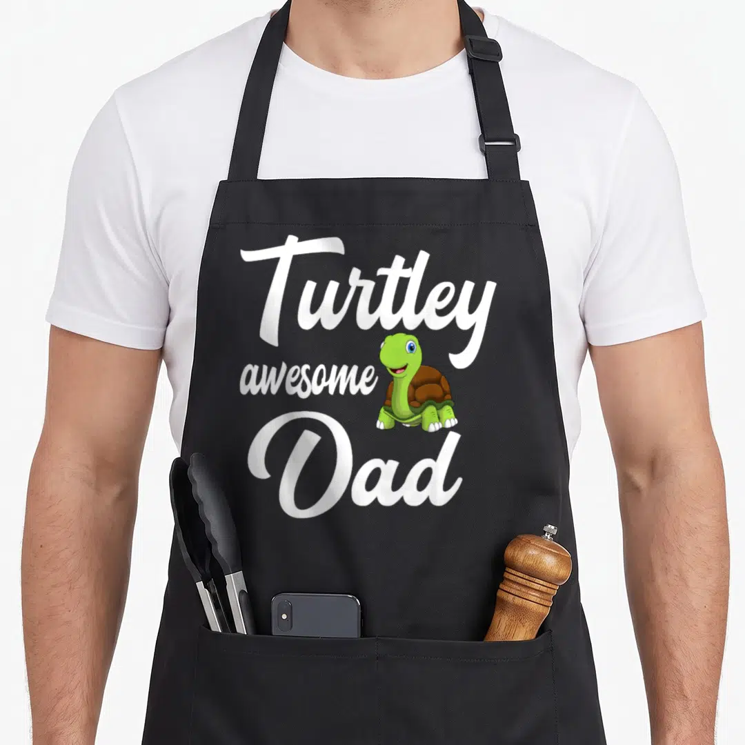 Turtley turtle dad apron - Cartoon green turtle with brown shell beside white text Turtley awesome Dad — shown on a black cotton canvas apron against a white background