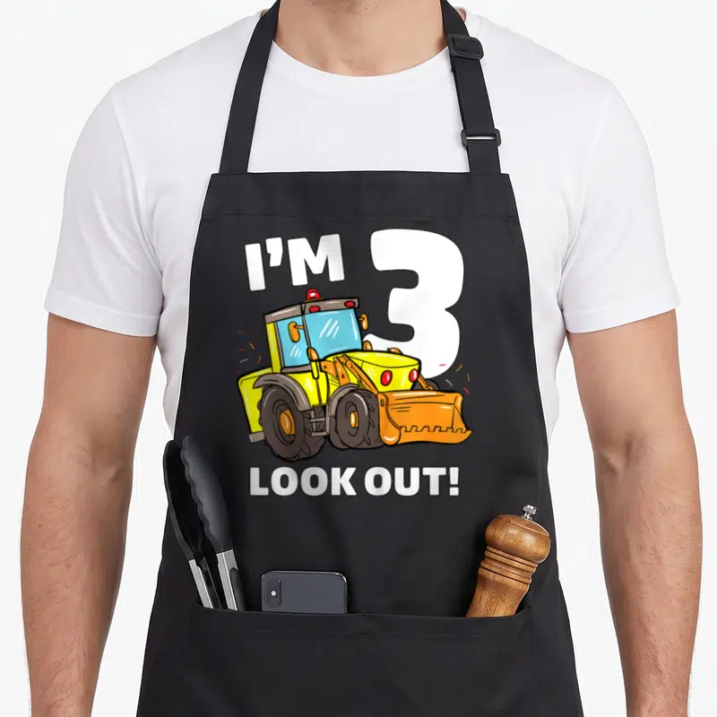 Three bulldozer truck apron - Cartoon yellow tractor with I'M 3 and LOOK OUT text in white, orange, and black — shown on a black cotton canvas apron against a white background