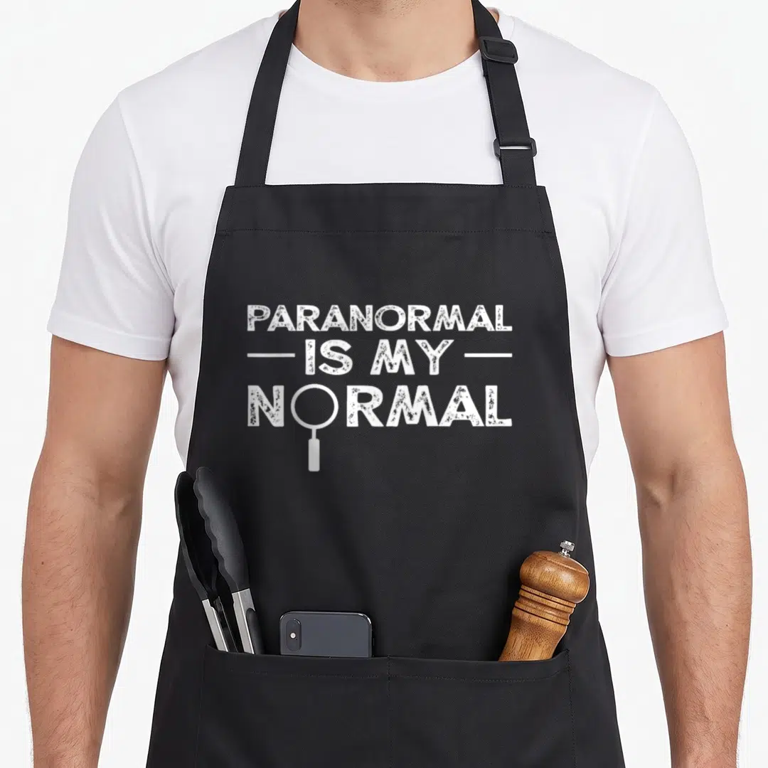 Paranormal investigator ghost apron - White distressed text reading Paranormal Is My Normal with a magnifying glass — shown on a black cotton canvas apron against a white background