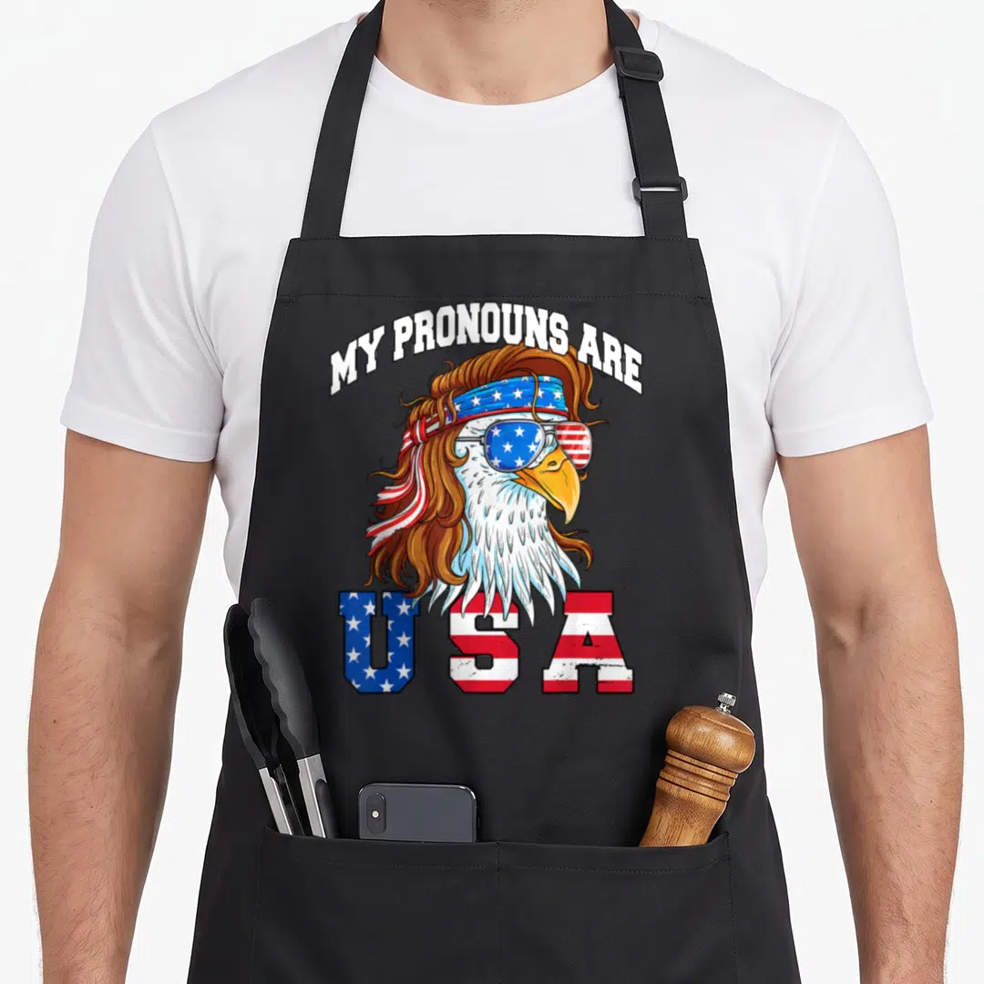 Pronoun 4th of july apron - Cartoon eagle with mullet and sunglasses wearing USA flag glasses and headband with arched — shown on a black cotton canvas apron against a white background