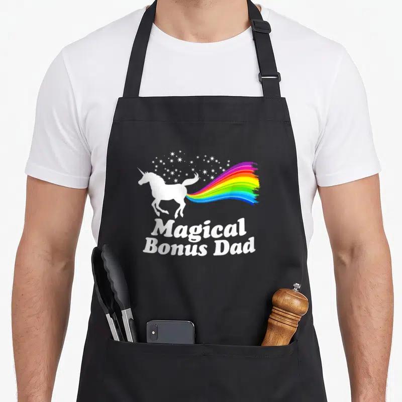 Magical unicorn apron - White unicorn with rainbow tail and Magical Bonus Dad text — shown on a black cotton canvas apron against a white background