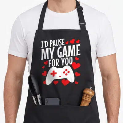 Gamer game valentine's day apron - Stacked white text above white controller surrounded by red hearts — shown on a black cotton canvas apron against a white background