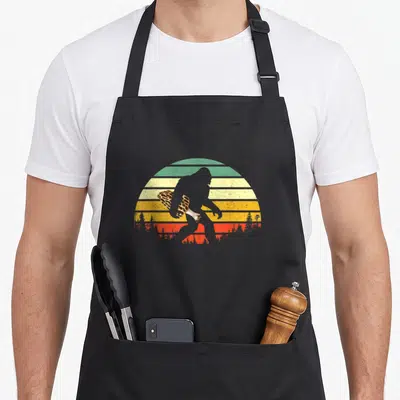 Sasquatch foraging apron - Retro striped sun with dark grey ape silhouette carrying a detailed morel mushroom and tree — shown on a black cotton canvas apron against a white background
