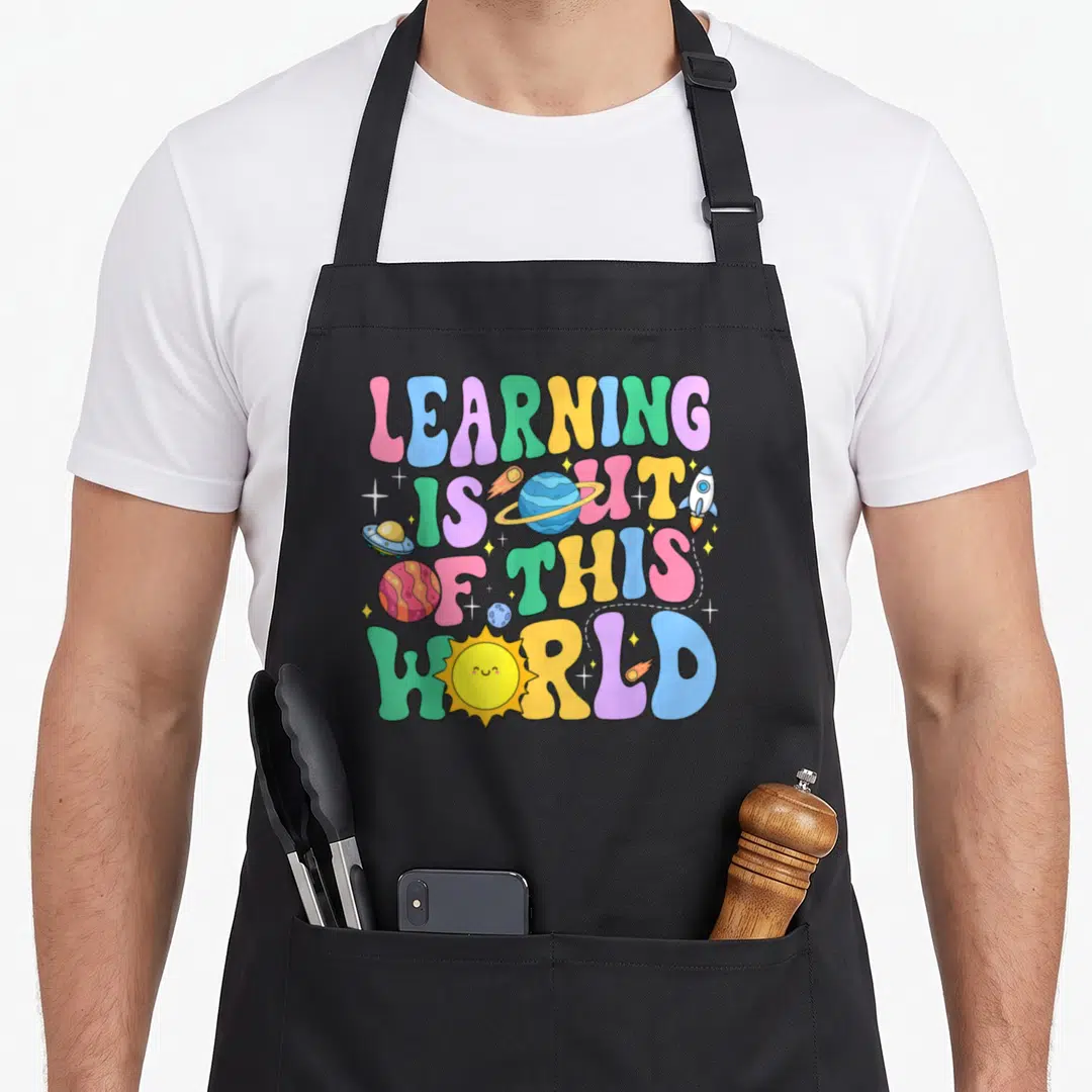 System teacher apron - Stacked colorful text reading LEARNING IS OUT OF THIS WORLD with cartoon planets and rocket — shown on a black cotton canvas apron against a white background