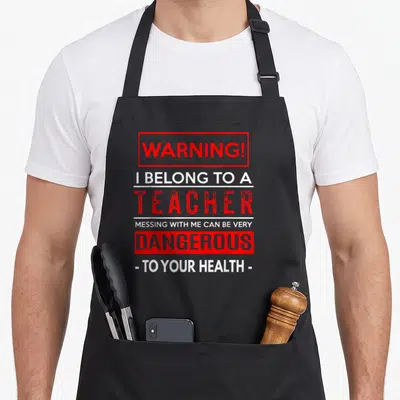 Wife teacher pocket apron - Red and white stacked warning text reading I belong to a teacher dangerous to your health — shown on a black cotton canvas apron against a white background