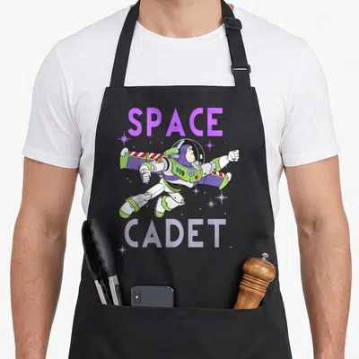 Cadet space apron - Buzz Lightyear cartoon flying between purple SPACE and CADET text with white stars — shown on a black cotton canvas apron against a white background