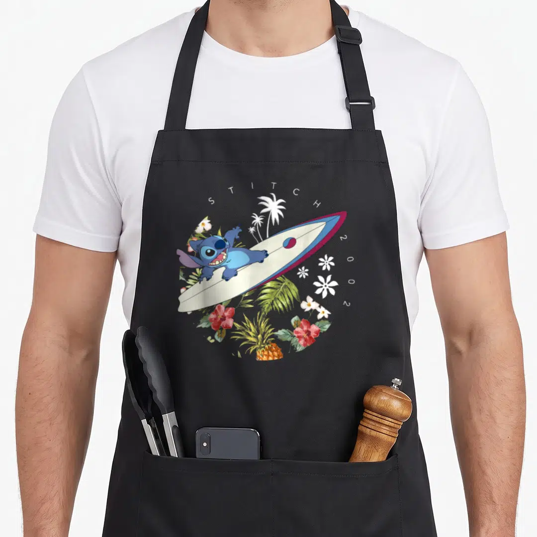 Surfing stitch apron - Blue Stitch lying on surfboard with STITCH 2002 text and tropical flowers — shown on a black cotton canvas apron against a white background