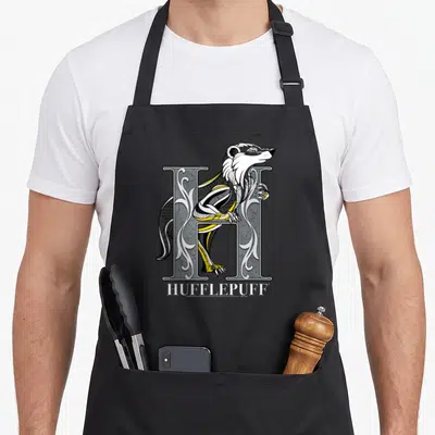 Mosaic badger apron - Badger entwined with ornate H letter and HUFFLEPUFF text in yellow and black line art — shown on a black cotton canvas apron against a white background