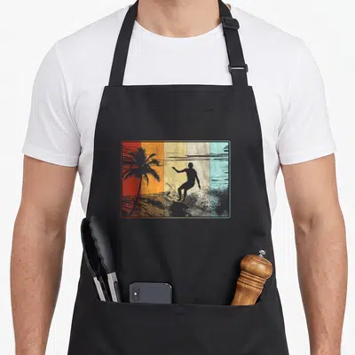 Beach surfing dad apron - Retro surfer silhouette on wave with palm tree on five colored vertical stripes enclosed — shown on a black cotton canvas apron against a white background
