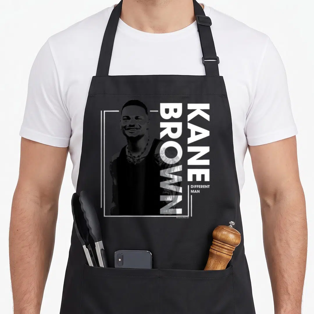 Kane line apron - Grayscale man photo left, vertical KANE BROWN DIFFERENT MAN text right, white L-border, two front — shown on a black cotton canvas apron against a white background