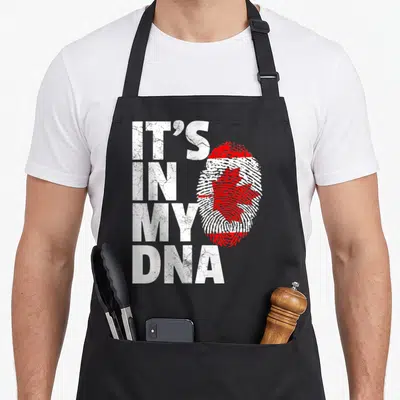 Canada pride apron - Distressed white IT'S IN MY DNA text beside a red maple leaf fingerprint graphic — shown on a black cotton canvas apron against a white background