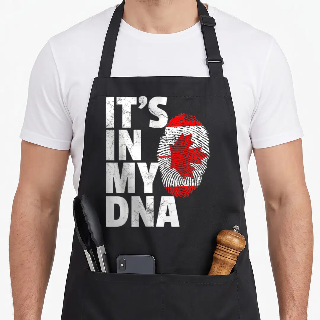 Canada pride apron - Distressed white IT'S IN MY DNA text beside a red maple leaf fingerprint graphic — shown on a black cotton canvas apron against a white background