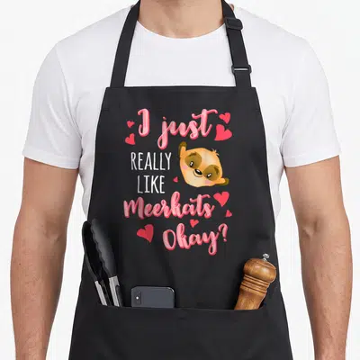 Okay animal apron - Cartoon meerkat head with pink hearts and text I just really like meerkats okay — shown on a black cotton canvas apron against a white background